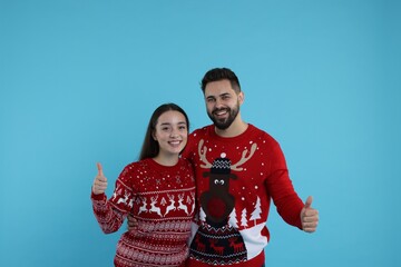 Wall Mural - Happy young couple in Christmas sweaters showing thumbs up on light blue background