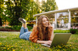 © maxbelchenko - Curly-haired woman in casual clothes with a laptop sits on the lawn on a sunny day. Happy freelancer woman working outdoors and enjoying the weather.