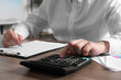 © New Africa - Man using calculator while taking notes at wooden table indoors, closeup