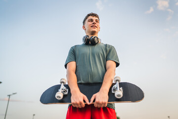 One man young adult caucasian teenager stand outdoor with skateboard on his shoulder and headphones posing at basketball court portrait happy confident wear shirt casual real person copy space