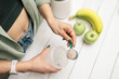 Young woman in jeans and shirt with measuring spoon in her hand puts portion of whey protein powder into shaker on white wooden table with bananas and apples. Process of making protein drink