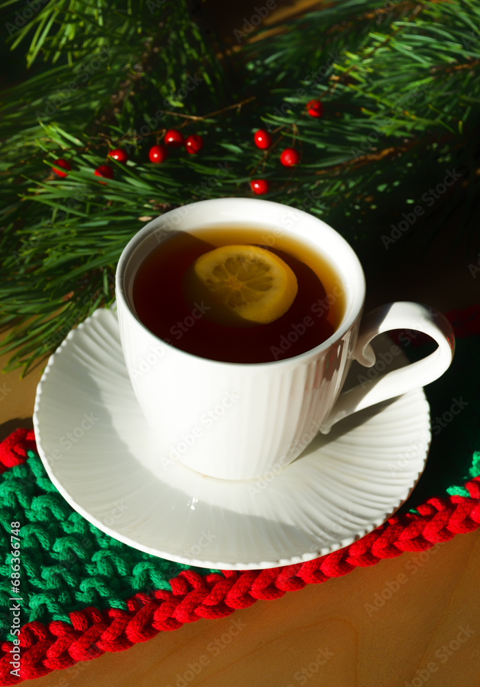 A cup of morning tea with lemon against the background of green Christmas tree branches. Decorations for the New Year and Christmas.