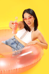 Wall Mural - Beautiful Asian woman with drink and inflatable ring on yellow background