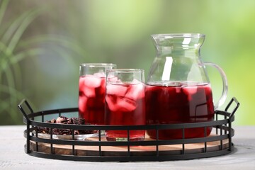 Wall Mural - Refreshing hibiscus tea with ice cubes and dry roselle flowers on white wooden table against blurred green background