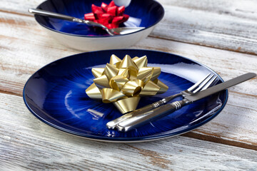 Wall Mural - Close up view of a holiday dinner table setting of a main dark blue dish and bowl with Christmas or New Years bow decorations plus cutlery on rustic white wood