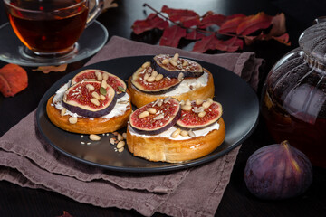 Wall Mural - Toast with cream cheese, fig slices, pine nuts, sunflower seeds and sesame seeds on a plate. Hot tea in a cup and teapot on a dark table