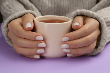 Wall Mural - Woman with white polish on nails holding cup of hot drink on violet background, closeup