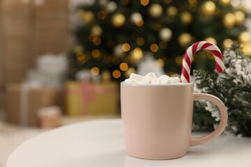 Canvas Print - Delicious cocoa with marshmallows and candy cane in cup against blurred Christmas lights, closeup. Space for text