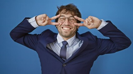 Poster - Joyful young man in business suit, smiling cheerfully, shows victory symbol with fingers on face over isolated blue background