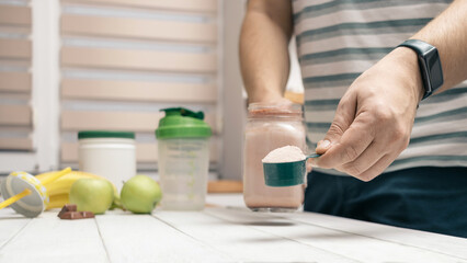 Canvas Print - Man holding measuring spoon with protein powder, glass jar of protein drink cocktail, milkshake or smoothie above white wooden table with chocolate pieces, bananas, apples