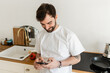 © LIGHTFIELD STUDIOS - Smiling and bearded tattooed man in white t-shirt using smartphone while standing near worktop