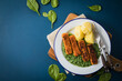 Panierte und gebratene Fischstäbchen mit Rahmspinat und Salzkartoffeln auf Emaille Teller mit Besteck, Holzbrett, Serviette, Spinat Blätter roh und dunkel blau Hintergrund