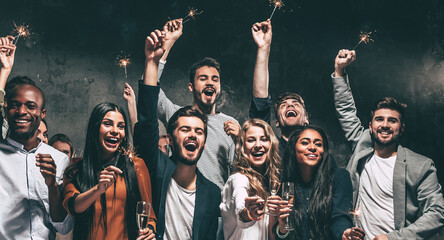 Wall Mural - Group of happy young friends holding champagne flutes and sparklers while having fun in night club together