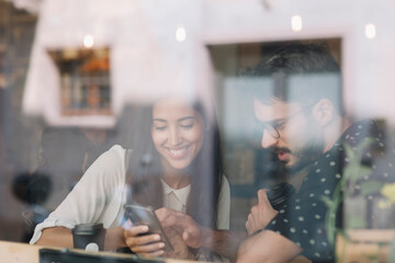 Wall Mural - Young couple with smartphone behind windowpane in a cafe