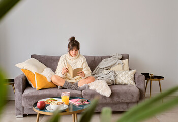 Wall Mural - Woman reading book while sitting on sofa against wall at home during winter