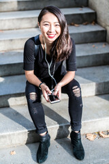 Poster - Portrait of young woman dressed in black sitting on steps listening music with earphones and cell phone