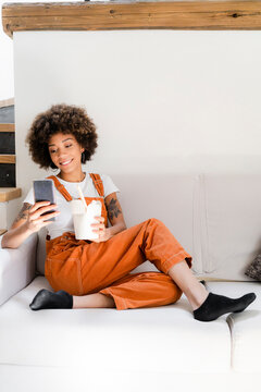 Portrait of smiling young woman sitting on the couch at home with box of Asian food looking at cell phone