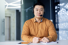 Portrait Of A Young Asian Male Businessman Sitting At A Desk In An Office Wearing A Headset And Looking Seriously At The Camera.