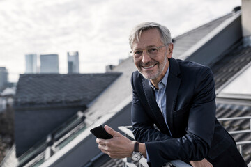 Wall Mural - Grey-haired businessman on balcony holding smartphone smiling into camera