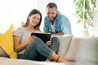 © tunedin - Happy couple at home in modern living room on couch looking at tablet together
