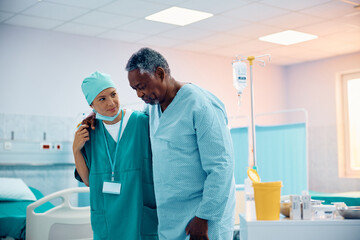 Wall Mural - Black senior man walking with help of nurse while recovering after surgery in hospital.