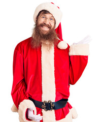 Sticker - Handsome young red head man with long beard wearing santa claus costume smiling cheerful presenting and pointing with palm of hand looking at the camera.