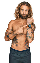 Handsome man with beard and long hair standing shirtless showing tattoos in hurry pointing to watch time, impatience, looking at the camera with relaxed expression