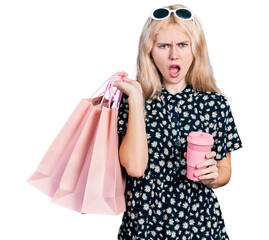 Canvas Print - Young caucasian woman holding shopping bags and coffee in shock face, looking skeptical and sarcastic, surprised with open mouth