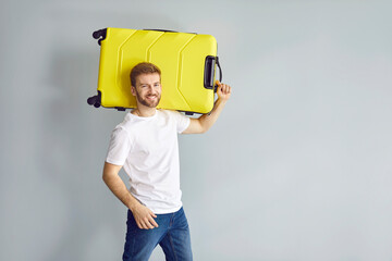 Portrait of a happy young man with a yellow suitcase. Handsome bearded guy in a tee shirt and jeans standing on a grey background and holding a suitcase on his shoulder. Travel, holiday trip concept