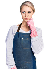 Sticker - Beautiful young blonde woman wearing cleaner apron and gloves serious face thinking about question with hand on chin, thoughtful about confusing idea