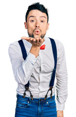 Canvas Print - Hispanic man with beard wearing hipster look with bow tie and suspenders looking at the camera blowing a kiss with hand on air being lovely and sexy. love expression.