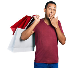 Wall Mural - Young african american man holding shopping bags covering mouth with hand, shocked and afraid for mistake. surprised expression