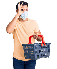 Sticker - Young hispanic man wearing shopping basket and medical mask smiling happy doing ok sign with hand on eye looking through fingers