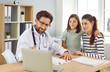 © Studio Romantic - Friendly family doctor talking to mother and child. Man pediatrician in white medical coat sitting at desk together with mum and daughter and showing diagnostic test results on modern laptop computer