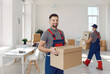 © Studio Romantic - Young happy smiling employee of moving service in overall standing in the living room of new house holding cardboard box and looking cheerful at camera with other mover on background.