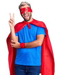 Wall Mural - Young blond man wearing super hero custome smiling with happy face winking at the camera doing victory sign. number two.