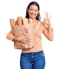 Sticker - Young beautiful latin girl holding paper bag with bread doing ok sign with fingers, smiling friendly gesturing excellent symbol