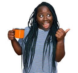 Wall Mural - Young black woman with braids drinking a cup coffee pointing thumb up to the side smiling happy with open mouth