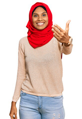 Wall Mural - Young african american woman wearing traditional islamic hijab scarf smiling friendly offering handshake as greeting and welcoming. successful business.