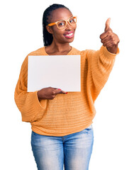 Sticker - Young african american woman holding cardboard banner with blank space smiling happy and positive, thumb up doing excellent and approval sign