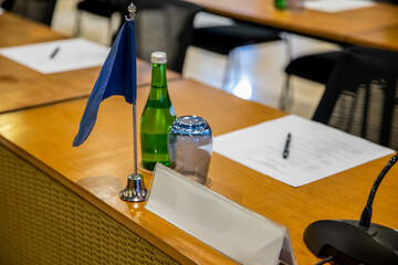 Gooseneck paging microphone on a table with blank name plate holder sign, unrecognizable flag, notes and water in conference hall during business meeting, summit, forum or other event