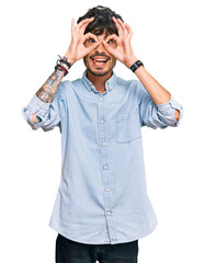 Sticker - Young hispanic man wearing casual clothes doing ok gesture like binoculars sticking tongue out, eyes looking through fingers. crazy expression.