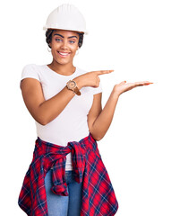 Sticker - Young african american woman with braids wearing hardhat and builder clothes amazed and smiling to the camera while presenting with hand and pointing with finger.