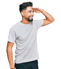 Canvas Print - Young man with beard wearing casual grey tshirt very happy and smiling looking far away with hand over head. searching concept.