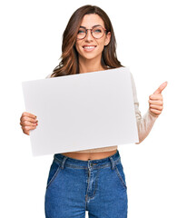 Canvas Print - Young brunette woman holding blank empty banner smiling happy and positive, thumb up doing excellent and approval sign