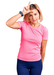 Wall Mural - Young blonde woman wearing sportswear making fun of people with fingers on forehead doing loser gesture mocking and insulting.