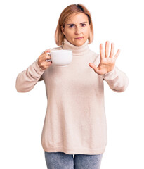 Sticker - Young blonde woman holding coffee with open hand doing stop sign with serious and confident expression, defense gesture