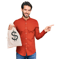 Sticker - Young hispanic man holding dollars bag smiling happy pointing with hand and finger to the side