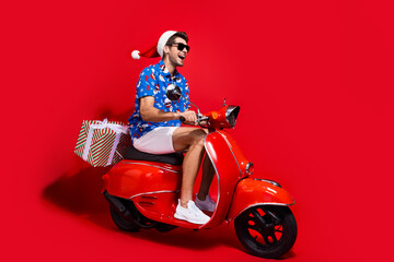 Sticker - Full body photo of handsome young guy deliver scooter presents dressed santa claus print x-mas outfit isolated on red color background