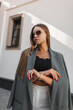 © alones - Fashion beautiful young business girl with vintage sunglasses in fashionable clothes with a stylish green blazer stands on the street near a white building. Pretty urban lady
